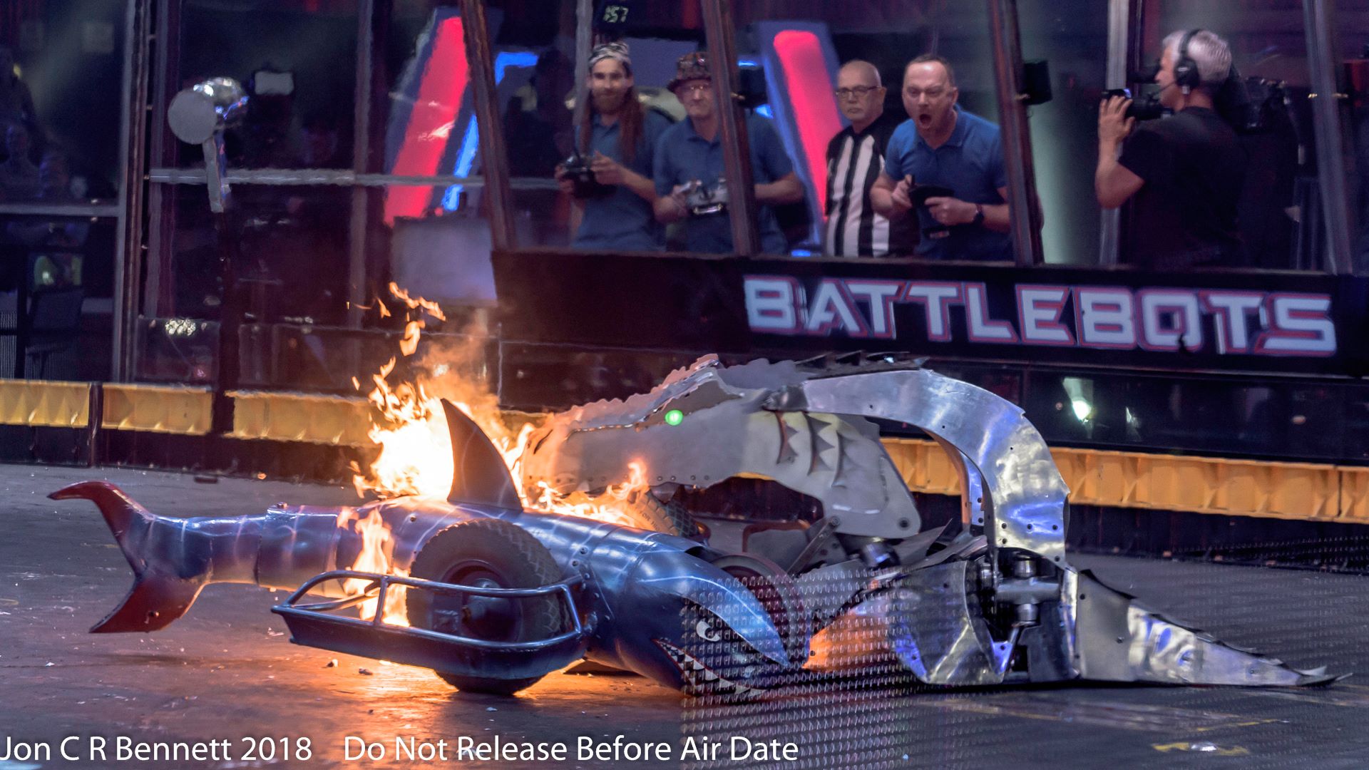 Battlebot arena featuring Sharkoprion battling another robot with flames shooting out of it's mouth and engineers in the background using remote controls